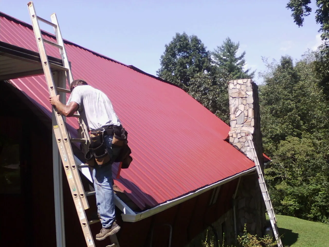 Roofer on a ladder beside a red metal roof and stone chimney — Soffit & Fascia Repair by Roofing Brothers in Laurel Bloomery